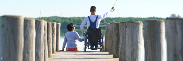 zwei Kinder, die Händchen haltend auf dem Steg sitzen, einer davon im Rollstuhl