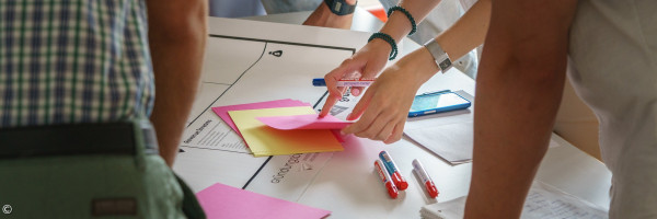 Menschen, die um einen Tisch stehen und mit Zetteln arbeiten